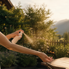 Frau spruht Mueckenspray auf ihren Arm auf sonniger Terrasse mit Garten und Bergen im Hintergrund